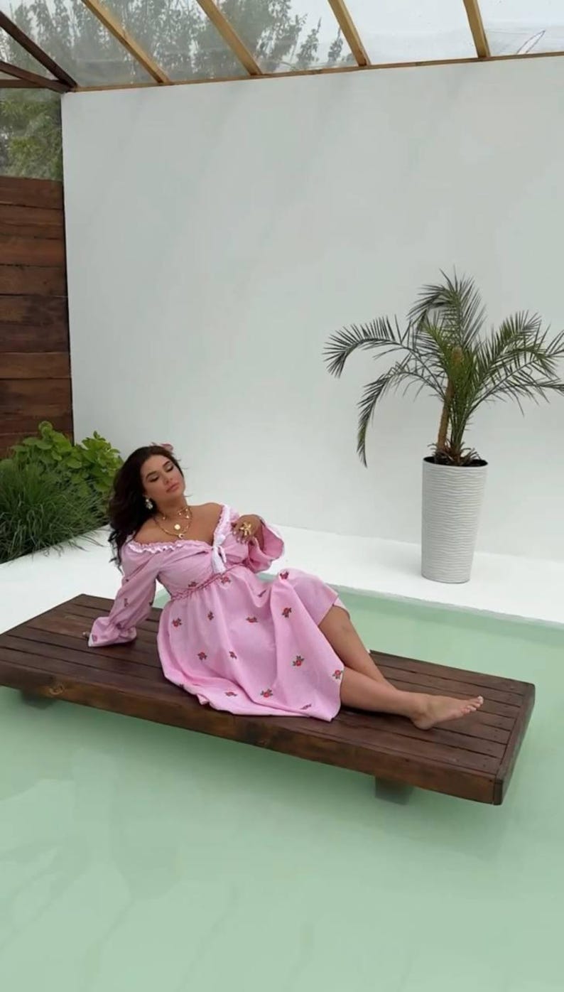 Puede incluir: Vestido rosa claro con estampado floral y mangas largas. El vestido tiene un ribete blanco y lo lleva una persona reclinada en una plataforma de madera. Una peque&ntilde;a palmera est&aacute; en el fondo.