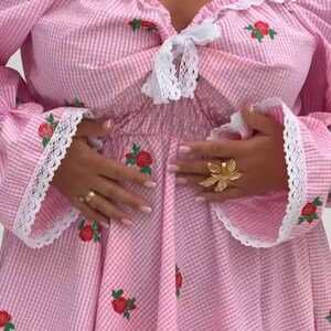 Puede incluir: Vestido de cuadros vichy rosa y blanco con bordado de rosas rojas. El vestido presenta un escote cuadrado, mangas largas con ribete de encaje y un lazo. Se aprecia un anillo de flores dorado y collares dorados superpuestos.
