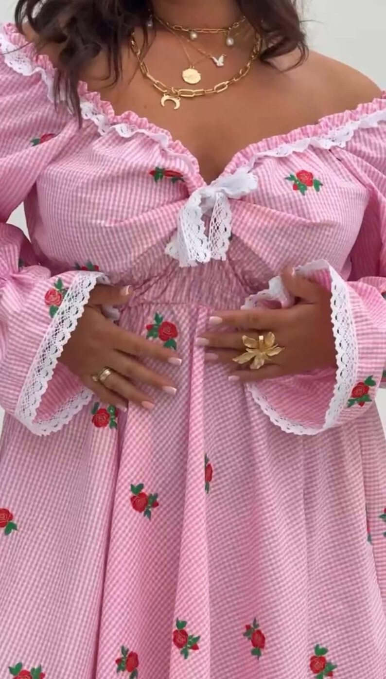 Puede incluir: Vestido de cuadros vichy rosa y blanco con bordado de rosas rojas. El vestido presenta un escote cuadrado, mangas largas con ribete de encaje y un lazo. Se aprecia un anillo de flores dorado y collares dorados superpuestos.