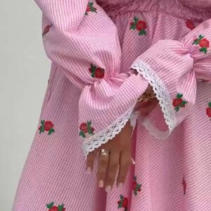 Puede incluir: Vestido a rayas rosas y blancas con rosas rojas bordadas. El vestido presenta mangas largas con detalles de encaje y un escote coraz&oacute;n. La tela tiene una textura seersucker, creando un estilo rom&aacute;ntico y femenino.