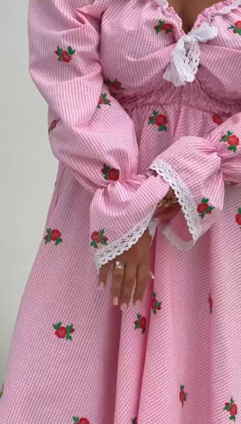 Puede incluir: Vestido a rayas rosas y blancas con rosas rojas bordadas. El vestido presenta mangas largas con detalles de encaje y un escote coraz&oacute;n. La tela tiene una textura seersucker, creando un estilo rom&aacute;ntico y femenino.