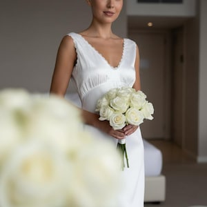 May include: A white satin wedding dress with a V-neckline and lace trim. The bride holds a bouquet of white roses. The dress is sleeveless and has a flowing skirt. The background is a neutral-toned room.