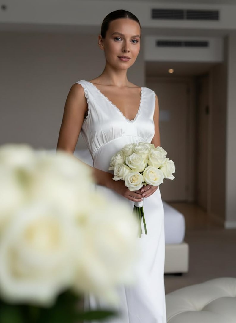 May include: A white satin wedding dress with a V-neckline and lace trim. The bride holds a bouquet of white roses. The dress is sleeveless and has a flowing skirt. The background is a neutral-toned room.