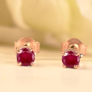 以下が含まれることがあります： ローズゴールドのスタッドピアス。それぞれに丸く深い赤色のルビーの宝石がセットされています。ピアスは柔らかくぼやけた背景に配置され、ルビーの鮮やかな色を際立たせています。