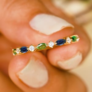Puede incluir: Un anillo de oro con piedras preciosas azules, verdes y transparentes alternadas. El anillo se sostiene entre los dedos, mostrando el delicado diseño y los colores vibrantes. Las piedras son de forma ovalada y están engastadas en una fina banda de oro.