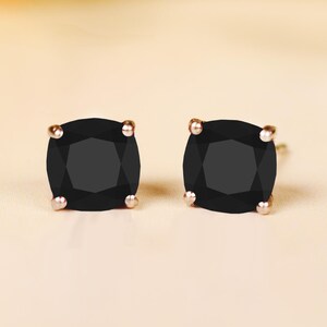 May include: Two black square-cut stud earrings with gold-coloured posts. The earrings are set against a soft, neutral background, highlighting the dark gemstones. A classic jewellery accessory.