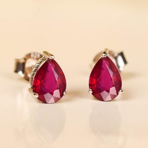 Könnte beinhalten: Ein Paar Ohrstecker mit tropfenförmigen, tiefroten Edelsteinen, gefasst in silberfarbenem Metall. Die Ohrringe werden auf einer reflektierenden Oberfläche präsentiert.