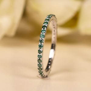 Puede incluir: Un anillo de plata con una banda de pequeñas piedras preciosas redondas de color verde esmeralda. El anillo se presenta sobre un fondo suave y neutro con acentos florales borrosos, resaltando su delicado diseño y color vibrante.