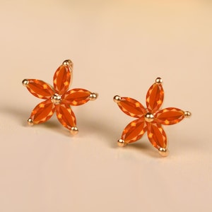 May include: A pair of gold-coloured flower stud earrings. Each earring has five orange petals around a central gold bead. The earrings are set against a neutral background.