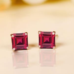 May include: A pair of square-cut, deep red gemstone stud earrings. The earrings are set in a silver metal, reflecting light. The gemstones are a vibrant, saturated color, and the earrings are displayed on a light-colored surface.