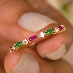 May include: A gold ring featuring alternating marquise-cut gemstones in green and red, interspersed with small, round, clear gemstones. The ring is held between fingers, showcasing its delicate design and vibrant colors.