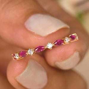 May include: A gold ring featuring alternating marquise-cut ruby gemstones and round diamonds. The ring's band is a thin, polished gold, and the gemstones are set closely together. The rubies are a deep red color, and the diamonds sparkle brightly.