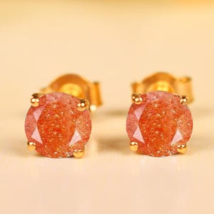 May include: A pair of gold stud earrings with round, faceted, orange-red gemstones. The earrings have a classic design with a gold setting that securely holds the stones. The gemstones have a sparkling, glittery appearance.