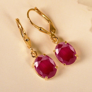 Puede incluir: Un par de pendientes dorados con cierre de palanca, cada uno con una gran gema roja oscura de forma ovalada. Los pendientes se colocan sobre un fondo suave y neutro, destacando el diseño elegante y el color de la joya.