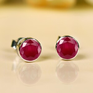 May include: A pair of stud earrings with round, faceted, deep red gemstones set in silver. The earrings are displayed on a light beige surface, with a soft, blurred background.