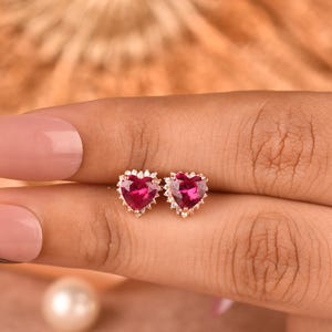 Puede incluir: Un par de pendientes de botón con piedras de color rosa intenso en forma de corazón engastadas en metal. Cada piedra central está rodeada por un halo de pequeñas piedras de acento claras y brillantes. Acabado metálico pulido y engaste clásico.