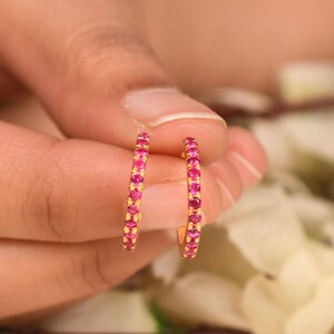 May include: A pair of small gold hoop earrings, each featuring a row of vibrant, round-cut pink gemstones set along the front-facing curve of the metal band, creating a bright and uniform appearance across the circular jewelry pieces.