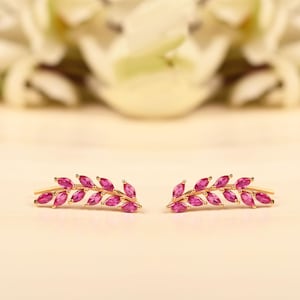 Może przedstawiać: Para złotych kolczyków w kształcie liści, każdy ozdobiony serią żywych różowych kamieni szlachetnych. Kolczyki są umieszczone na miękkim, neutralnym tle, z rozmytymi białymi kwiatami w tle.