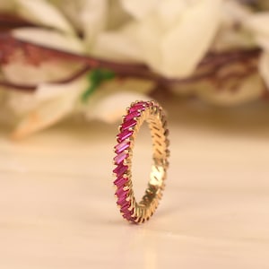 May include: A gold eternity band featuring a continuous row of vibrant, deep pink baguette-cut stones set vertically around the entire circumference of the ring, creating a uniform and structured design with a polished metallic finish.
