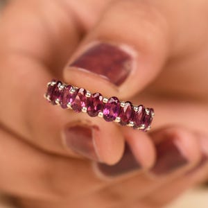 May include: A silver ring featuring a row of oval-cut, deep pink gemstones. The gemstones are set closely together, creating a continuous band of color. The ring is held against a blurred background.