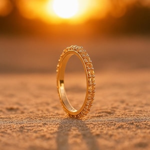 May include: A gold ring, set with small, round gemstones, sits on sand. The background is blurred, showing a bright sunset. The ring's shadow is cast on the sand. The ring is likely made of gold.