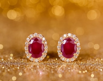 Peut inclure: Paire de boucles d'oreilles à clous en or, ornées de rubis ovales entourés de diamants. Les boucles d'oreilles sont posées sur une surface dorée scintillante, avec un fond flou.