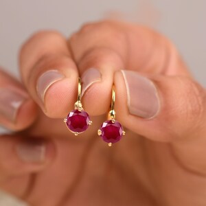 May include: A pair of gold leverback earrings, each featuring a single round, faceted deep red gemstone set in a four-prong setting. The earrings have a polished gold finish and a classic drop design that hangs securely from the ear wire mechanism.
