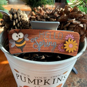Puede incluir: Pinza para el pelo de madera con una abeja y flores, y el texto "Hello Spring".