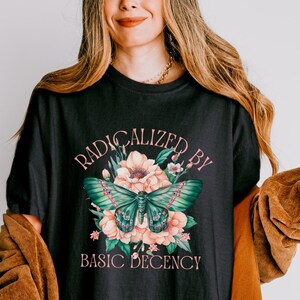May include: Black t-shirt with a floral and butterfly design. The graphic includes the words "RADICALIZED BY BASIC DECENCY" in a pink script. The butterfly is teal and the flowers are pink and cream.