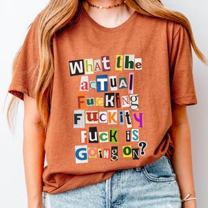 May include: A rust-colored t-shirt with the text "What the actual fucking fuckity fuck is going on?" in a ransom note style. The shirt is paired with light blue denim jeans.
