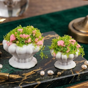 May include: Two white ceramic urns filled with green and pink floral arrangements sit on a black and green marble surface. A gold-tipped fountain pen, pearls, and a vintage lamp are also visible.