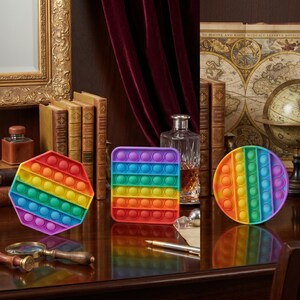 May include: Three colorful pop-it fidget toys in rainbow hues. The shapes include a hexagon, a square, and a circle. The toys are on a dark wooden surface with books, a framed picture, and a globe in the background.