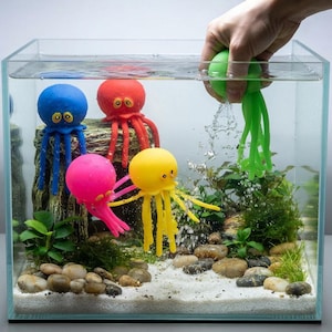 May include: Four colorful octopus-shaped toys in a glass aquarium. One blue, one red, one pink, and one yellow octopus are positioned among rocks and plants, while a green octopus toy is being submerged into the water by a hand.