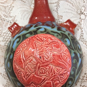 May include: A handcrafted ceramic bottle with a red neck and accents. The body is dark with blue and green swirls, and a raised red circular design. Two red, square-shaped handles are on the sides.