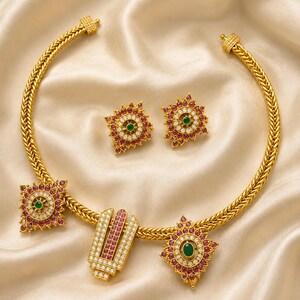 May include: A gold necklace featuring a braided chain with two square floral-shaped pendants and a central U-shaped pendant, all set with red and white stones and a green center stone, accompanied by two matching square floral-shaped stud earrings.