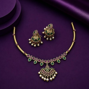Puede incluir: Un conjunto de collar y pendientes jhumka en tono dorado con piedras redondas y en forma de lágrima de color verde y magenta. El collar tiene una cadena de metal trenzado y un colgante central con cuentas blancas colgantes, y los pendientes son de cúpula.