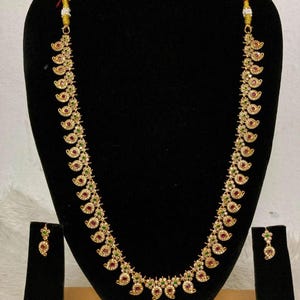 Pode incluir: Um colar longo em tom dourado com um motivo de caxemira repetitivo adornado com pequenas pedras vermelhas e verdes e detalhes brancos. O conjunto inclui um par de brincos a condizer, cada um com um design de caxemira e pedras.