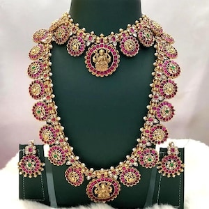 May include: A gold-toned necklace and earring set featuring circular motifs with deep pink stones, small green accents, and central gold deity figures. The necklace has a layered design with ornate floral-like patterns and intricate metalwork.
