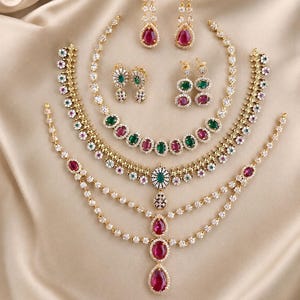 May include: A jewelry set featuring three gold-tone necklaces and three pairs of earrings. The pieces are adorned with clear white stones and alternating oval-shaped red and green gemstones. One necklace includes a multi-layered chain design.