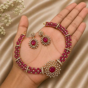 May include: A gold-toned jewelry set consisting of a necklace and a pair of matching earrings. The necklace features three rows of small, round, deep pink stones accented by clear white crystals, with a large circular floral-shaped pendant.
