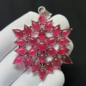 Puede incluir: Un colgante de tono plateado con una piedra preciosa circular central rodeada por múltiples piedras rosas en forma de lágrima dispuestas en un patrón de estrella. El engaste metálico tiene un acabado texturizado y oxidado, con una anilla triangular.