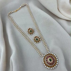 May include: A gold-toned necklace featuring three strands of small white pearls and a large circular pendant with a central green stone surrounded by magenta stones and pearls, accompanied by two matching circular stud earrings with green stones.