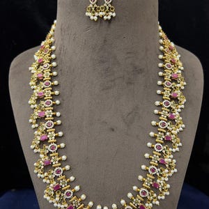 May include: A gold-toned necklace and matching earring set featuring rectangular pink stones, clear accent crystals, and small white pearl-like beads. The necklace has a long, intricate chain design with repeating floral and geometric motifs.