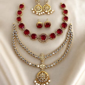 Puede incluir: Un conjunto de joyería que incluye un collar dorado con piedras rojas cuadradas, un tocado de dos niveles con cristales transparentes y un colgante floral circular, pendientes colgantes con perlas y un par de pendientes de botón rojos.