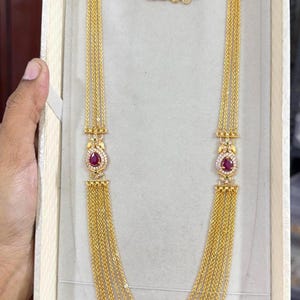 May include: A long gold necklace featuring multiple delicate chains that drape in layers. The design includes two symmetrical decorative connectors, each set with a central teardrop-shaped red stone surrounded by small white stones.