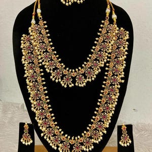 Può includere: Un set di gioielli color oro composto da una lunga collana a strati e due paia di orecchini pendenti abbinati. I pezzi sono decorati con piccole perle bianche e pietre rosse e verdi a forma di goccia con motivi floreali.