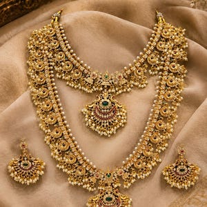 May include: A gold-toned jewelry set consisting of two layered necklaces and a pair of matching earrings. Each piece features intricate metalwork, small white beads, and central green and red stone accents in a crescent-shaped pendant design.