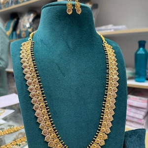 May include: A gold-toned long necklace featuring a series of circular, coin-like pendants with intricate embossed detailing and small red and green stone accents, accompanied by a matching pair of small, dangling stud earrings on a display.