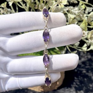 May include: A silver bracelet featuring four marquise-cut purple amethyst gemstones set in metal prongs. The stones are arranged in a vertical line, connected by small silver circular links, creating a delicate and symmetrical jewelry piece.