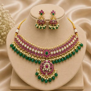 May include: A gold-toned necklace and matching jhumka earring set featuring deep pink and green stones, clear white crystals, and small white pearls. The necklace has a central circular pendant and dangling green beads along the bottom edge.